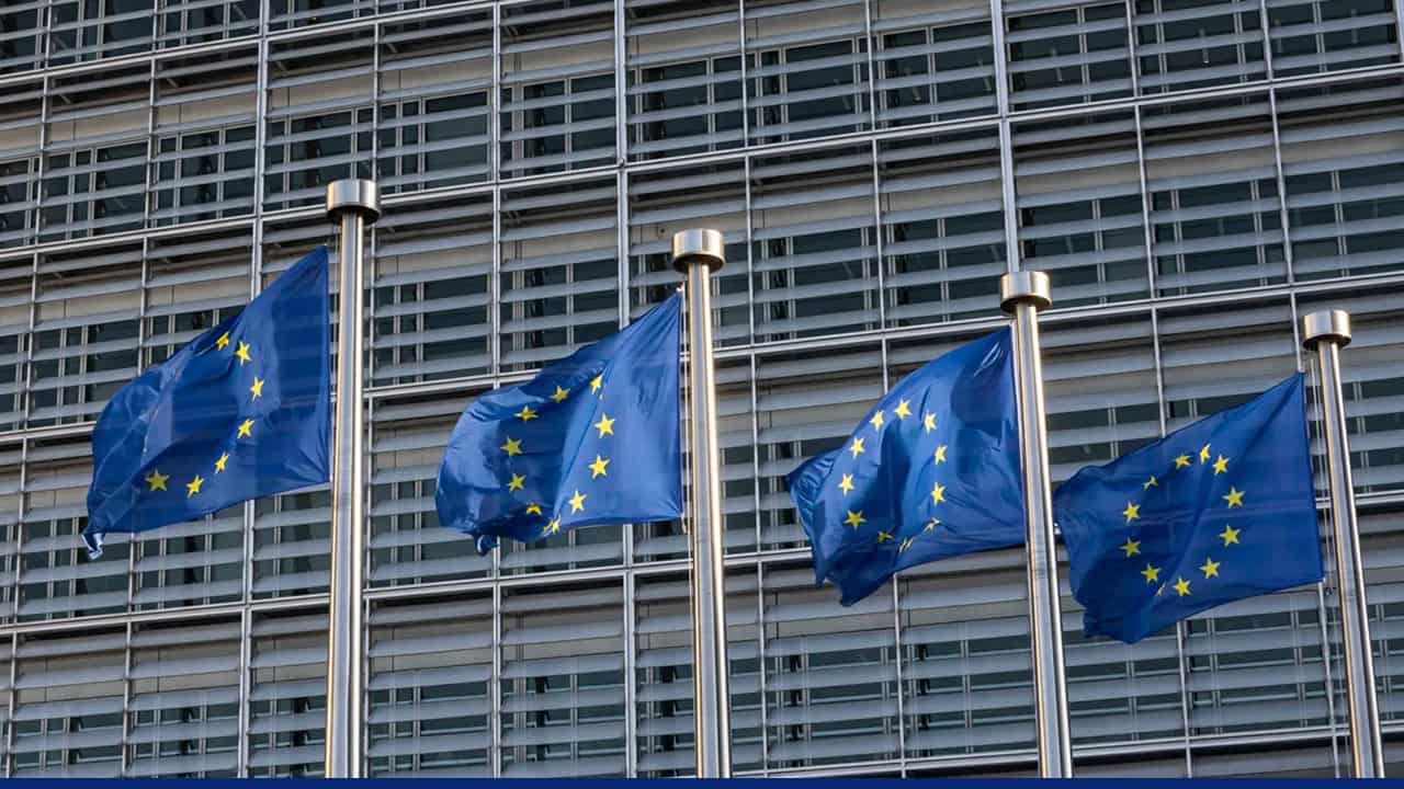 European Union flags flying outside a modern glass building, symbolizing EU institutions and governance.
