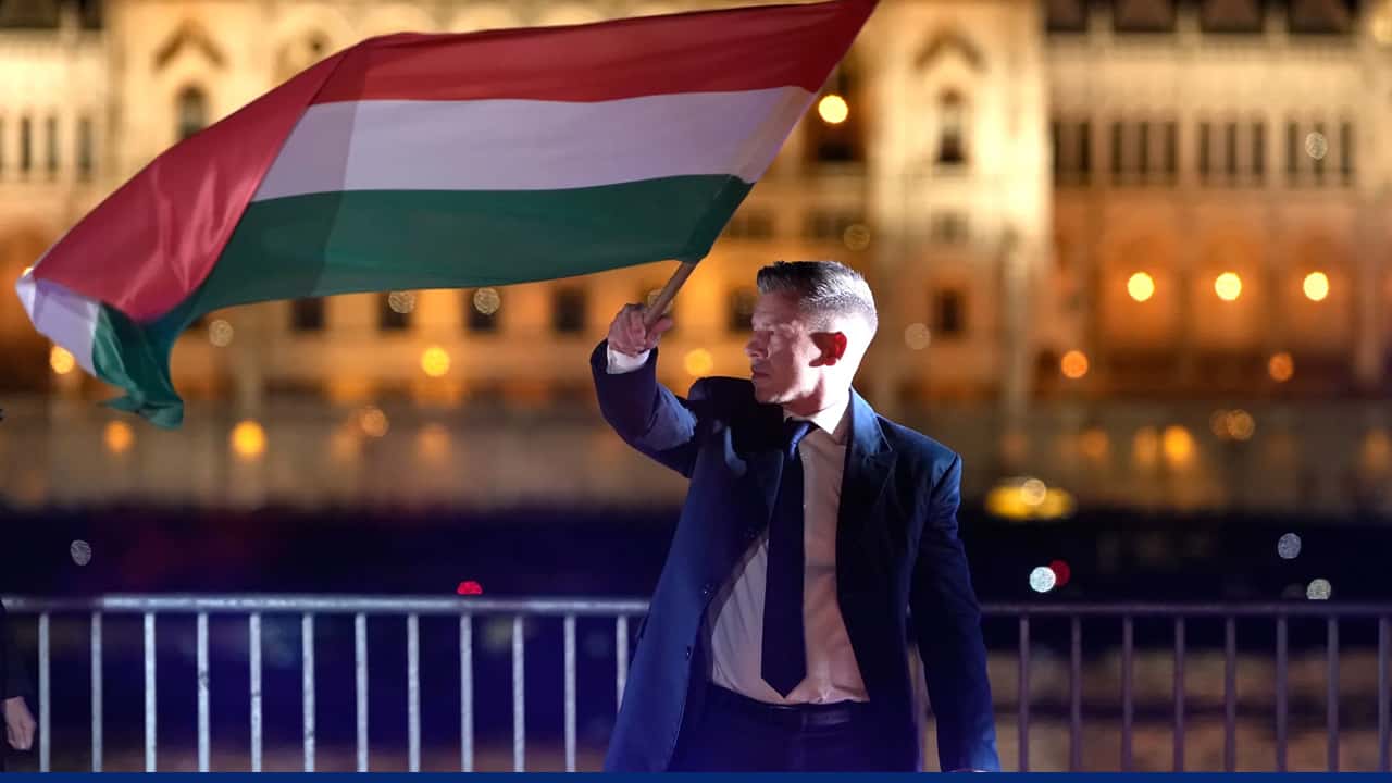 A man in a suit waves the national flag of Hungary (red, white, and green horizontal stripes) at night, standing by a riverside railing with illuminated historic buildings in the background.