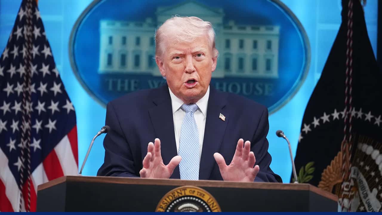A man speaks at a podium during a press briefing, with U.S. flags and a White House backdrop behind him.