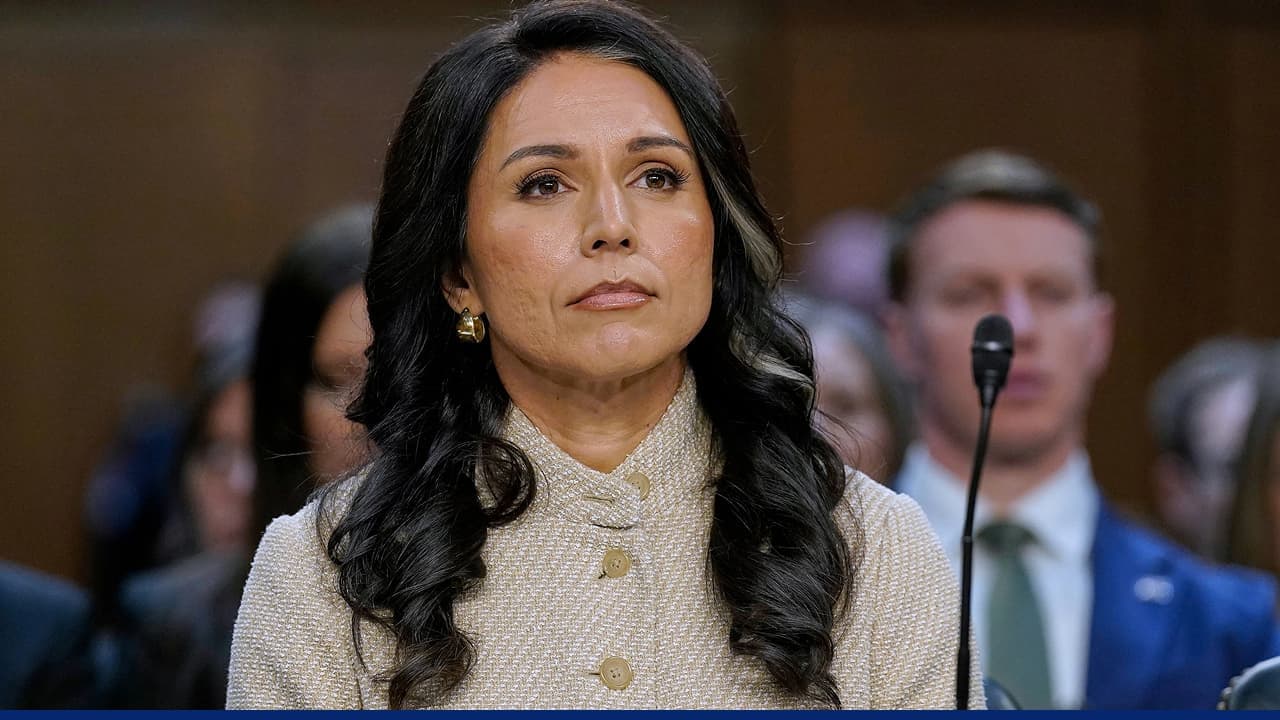 Tulsi Gabbard testifies during a congressional intelligence hearing, looking serious and composed while seated before a microphone, with officials blurred in the background.