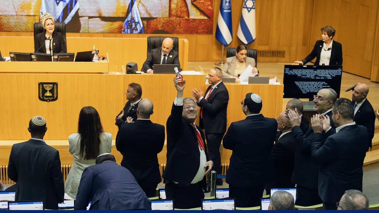 Lawmakers inside a parliamentary chamber with Israeli flags visible, as one man raises an object while others stand and applaud, and officials sit at the front desk.